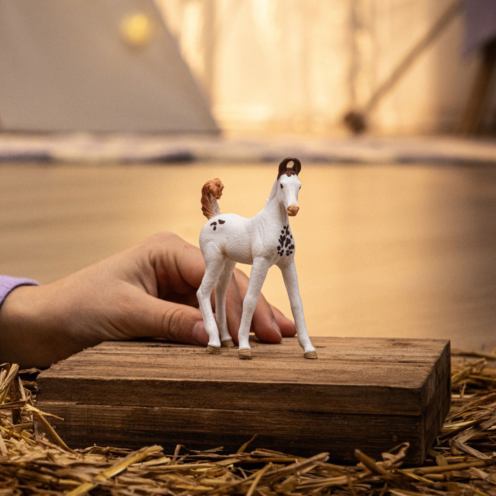 A white foal figurine with brown spots stands on a wooden block surrounded by hay, with a hand behind it