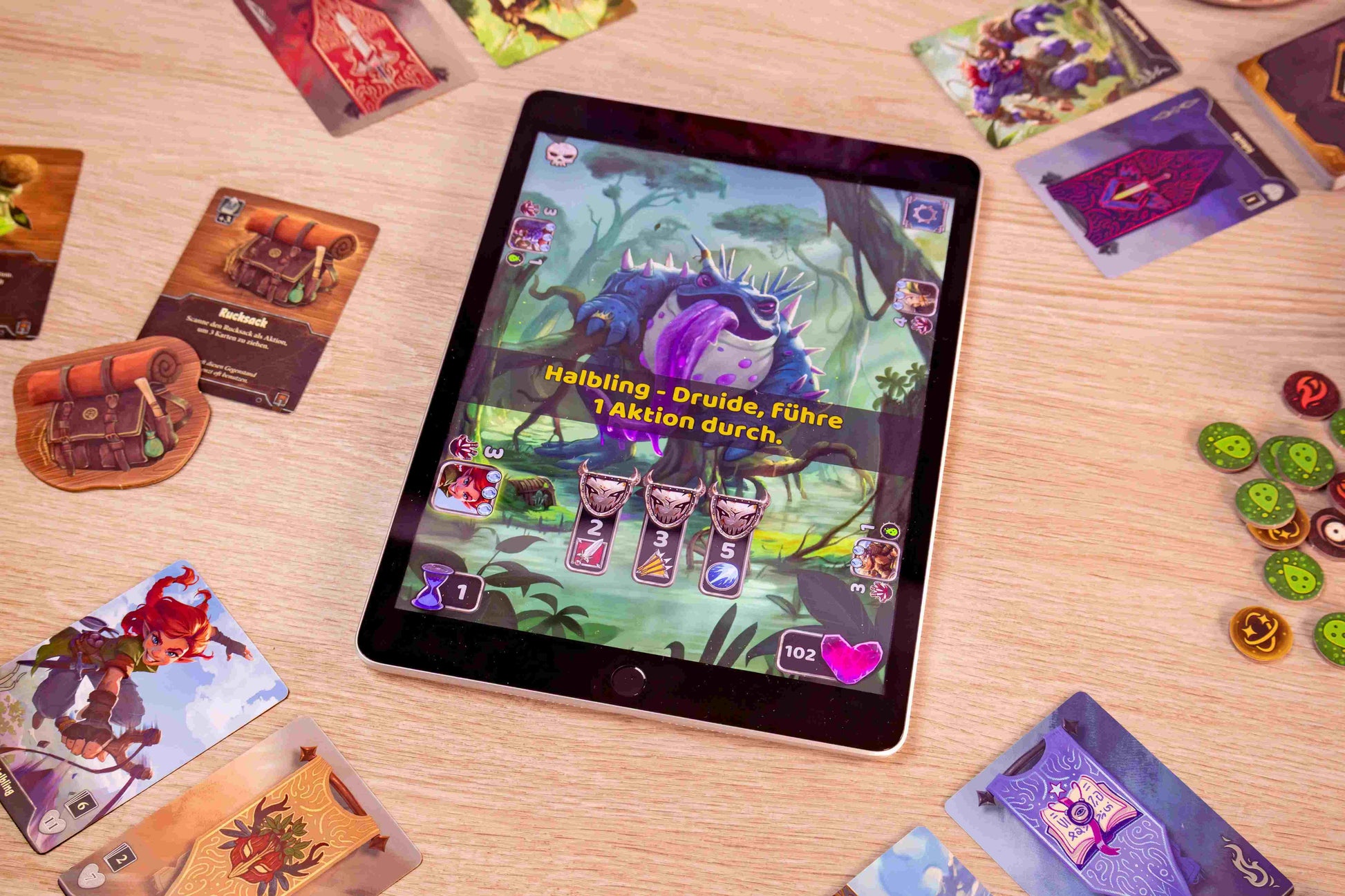 An overhead view of a tablet displaying the Boss Fighters game with a purple Halbling-Druid boss, surrounded by various illustrated physical cards and circular tokens on a wooden surface