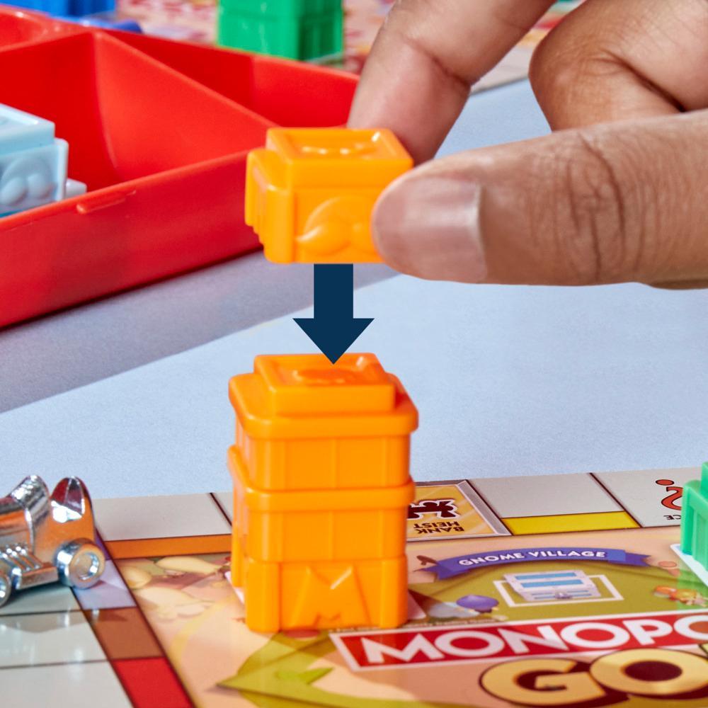 A hand places an orange plastic chest piece onto a stack of two others on a Monopoly GO! game board, with a blue arrow indicating the downward movement