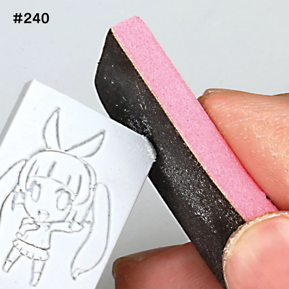 A hand uses a pink and black sanding block to smooth the edge of a white block carved with an anime character