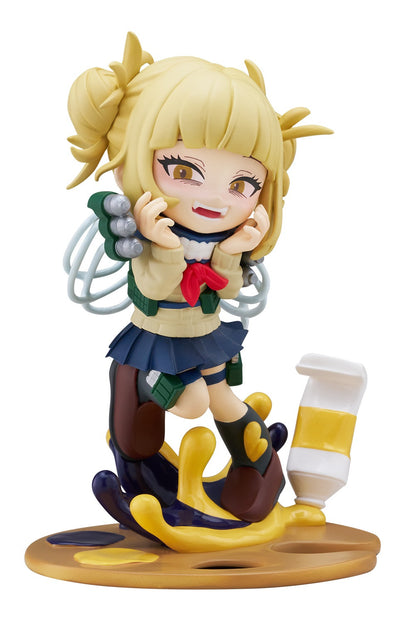 Chibi anime figure of a blonde girl with fangs, hands near face, in uniform on an artist's palette with paint and tube
