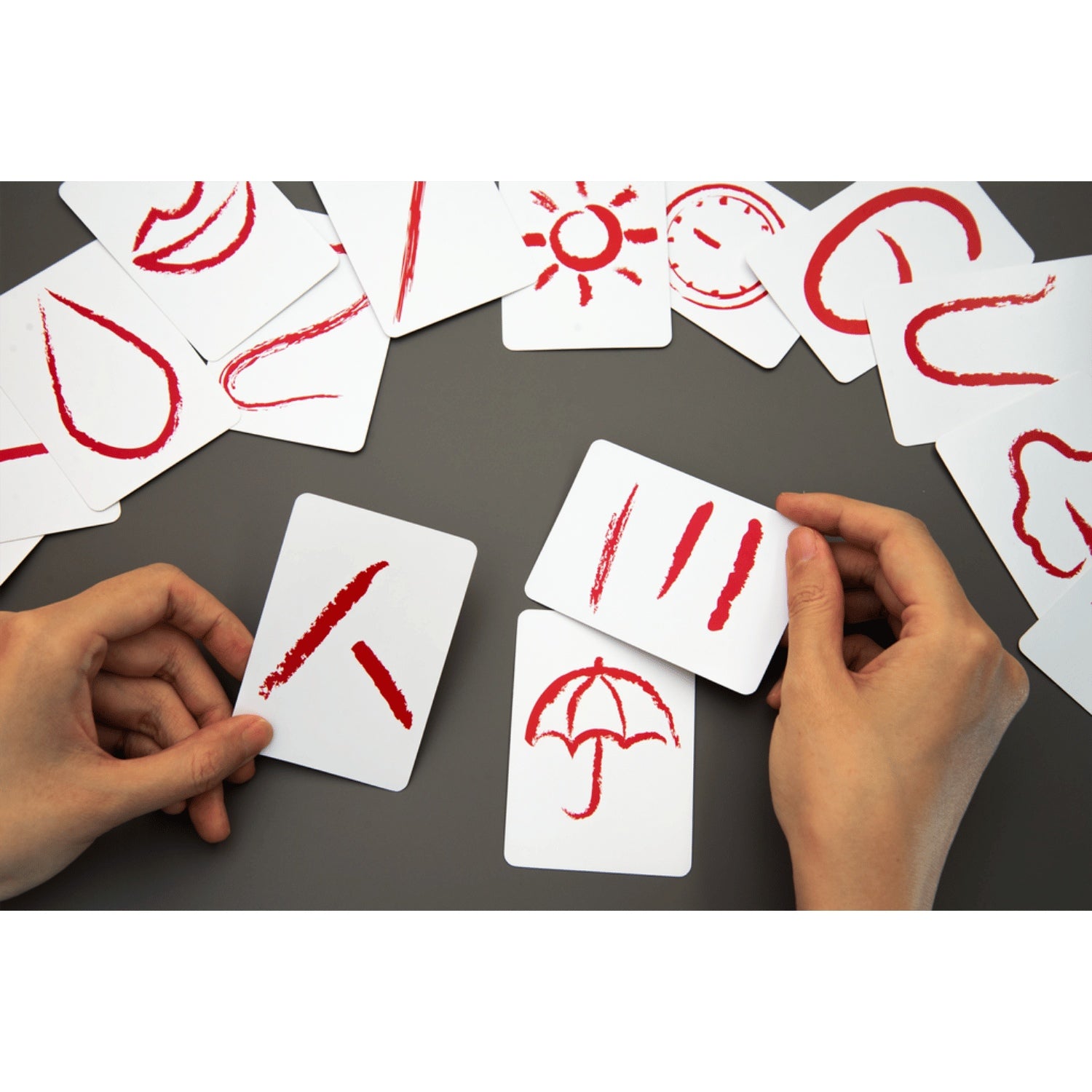 Two hands hold Oink Games Dying Message cards with red brushstroke symbols while other cards showing symbols like a sun, clock, and umbrella are scattered on a dark surface