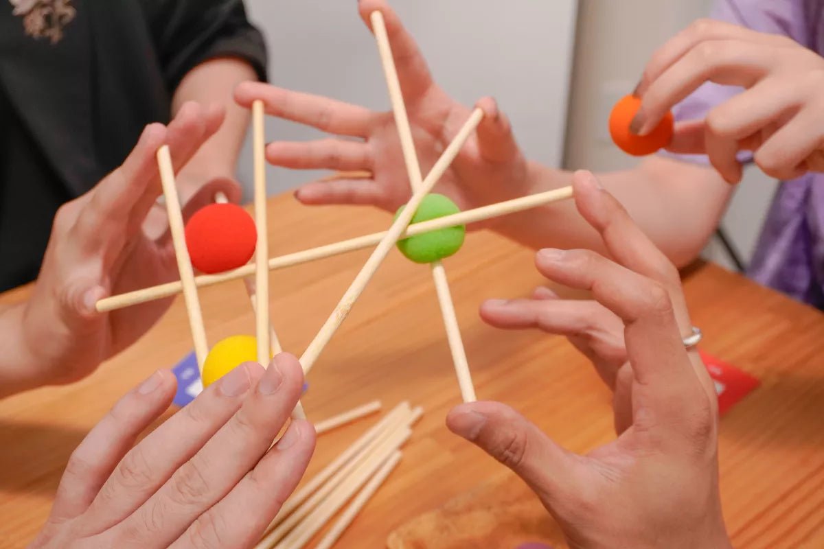 Hands play a Yubibo 4580439940659 game carefully adding wooden sticks and colorful foam balls red yellow green orange to a growing structure