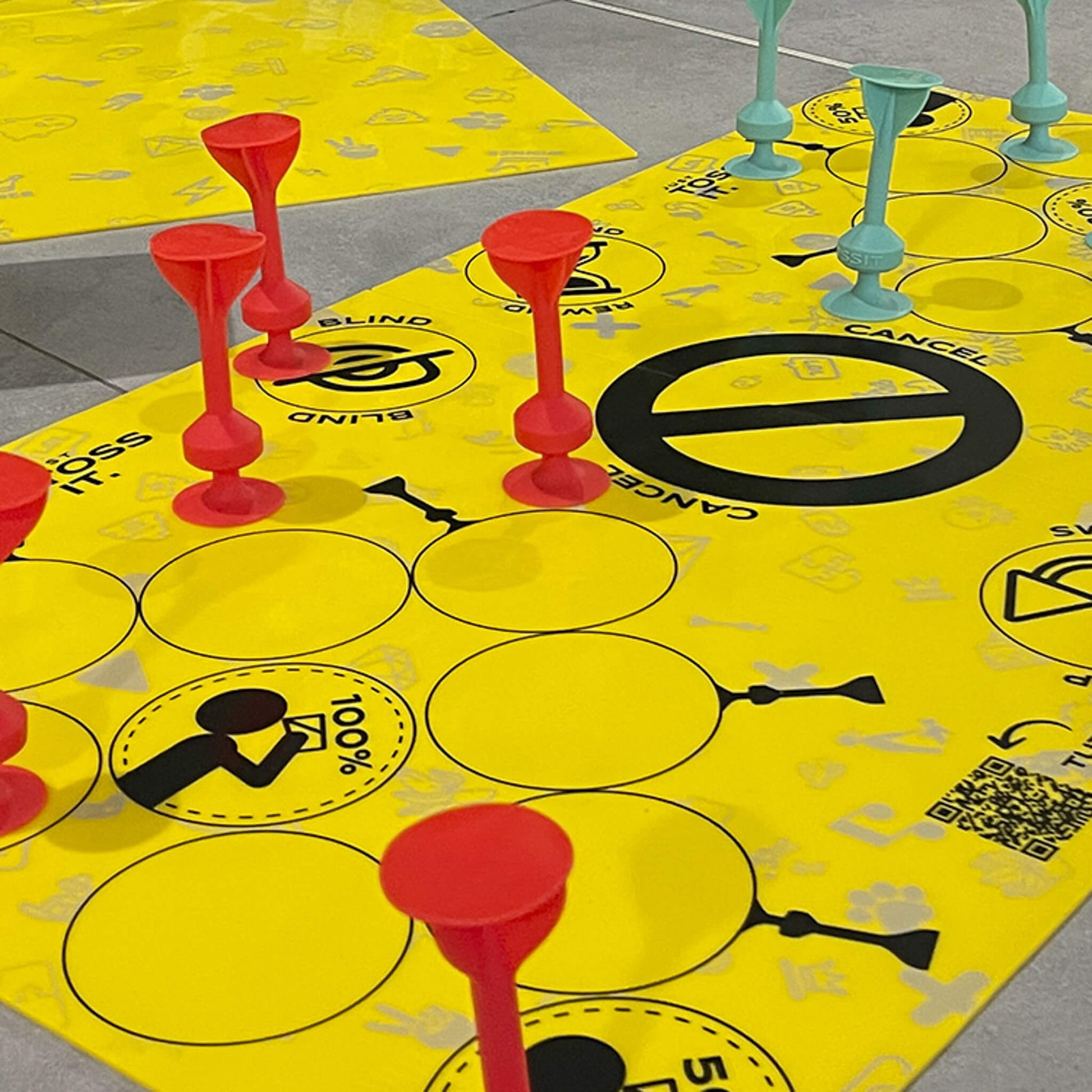 Bright yellow Tossit game board featuring black instructions and target circles, currently holding multiple tall red and teal plastic cup-shaped playing pieces
