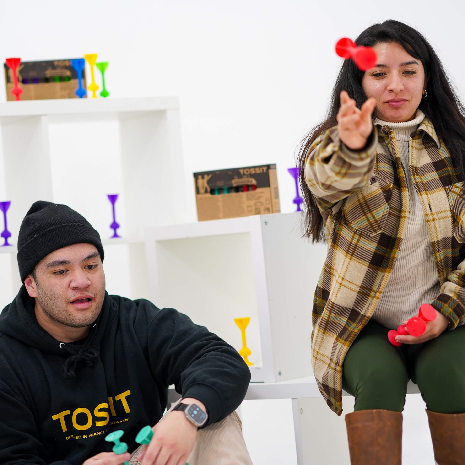 Two adults enjoy playing the Tossit Starter Pack game, featuring multicolored silicone suction-cup darts being thrown and displayed on white shelving