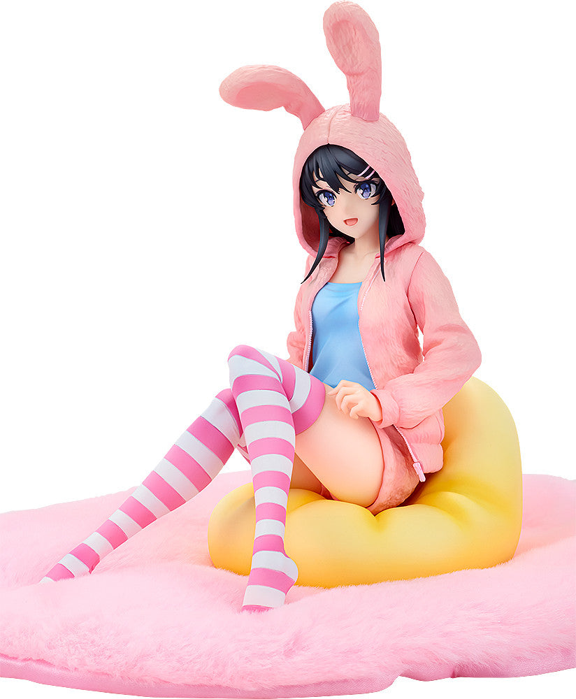 Anime figure in pink bunny hoodie and striped socks sits on a yellow cushion on a pink fluffy surface