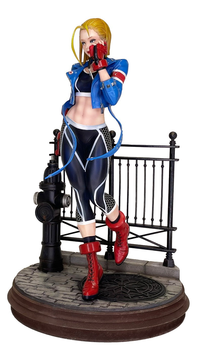 Side view of Street Fighter 6 Cammy figure in blue jacket, black crop top, red boots, patterned leggings, leaning on a fire hydrant and fence on a cobblestone base