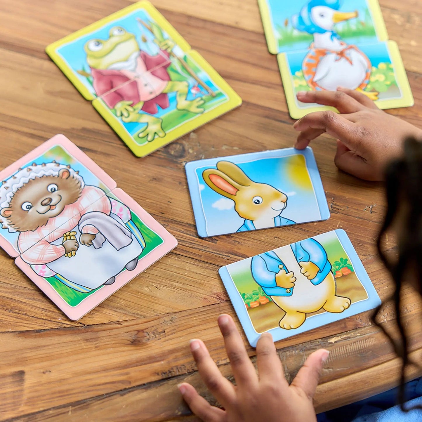 Child's hands assemble Peter Rabbit Heads and Tails game cards on a wooden table, showing cartoon animal characters like a frog, duck, hedgehog, and Peter Rabbit