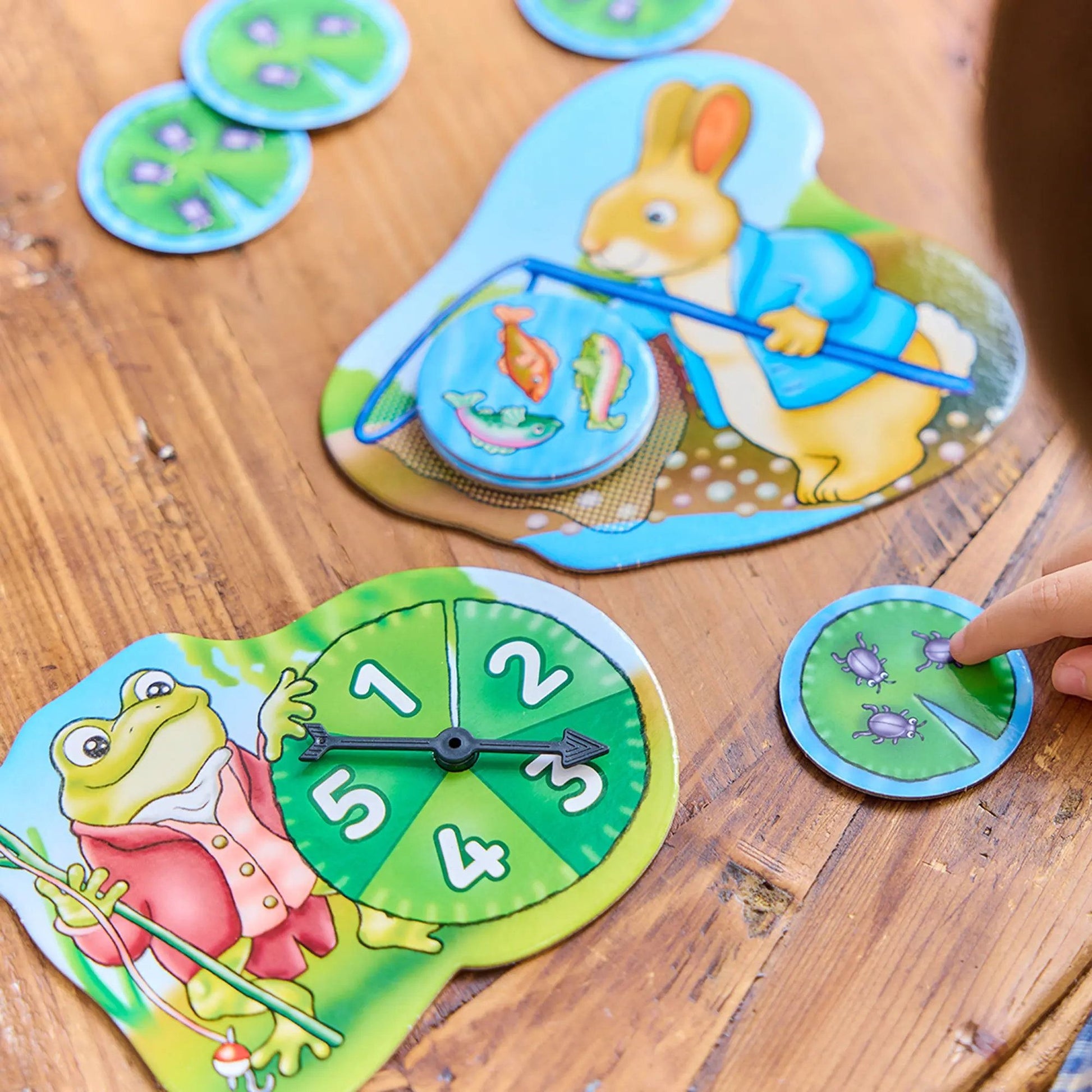 Orchard Peter Rabbit - Fish And Count game pieces on a wooden table including Peter Rabbit and frog boards, a number spinner, and a child's hand on a lily pad token