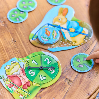 Orchard Peter Rabbit - Fish And Count game pieces on a wooden table including Peter Rabbit and frog boards, a number spinner, and a child's hand on a lily pad token