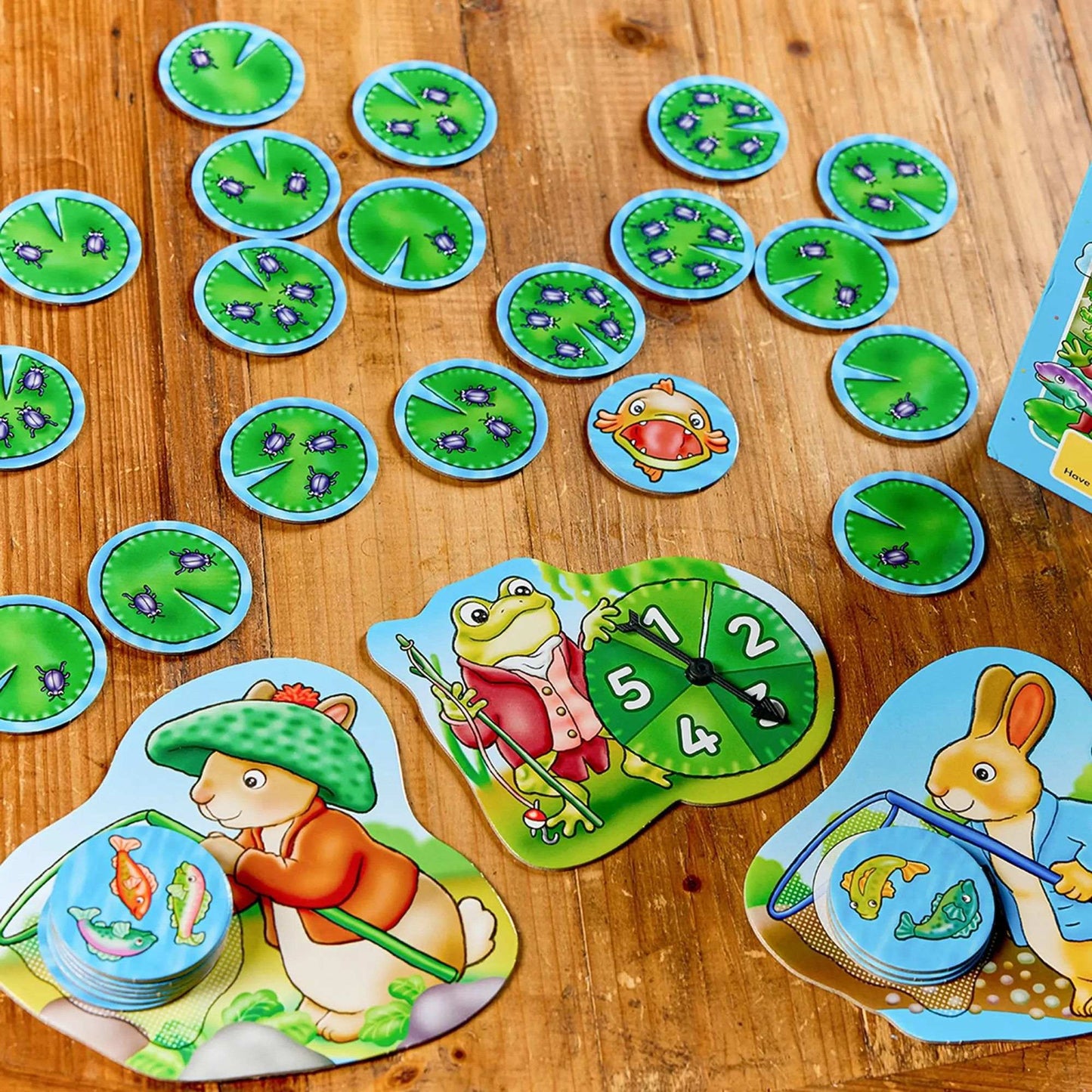 Orchard Peter Rabbit - Fish And Count game pieces including lily pads with bugs, a frog with a spinner, and rabbits fishing for fish tokens on a wooden table