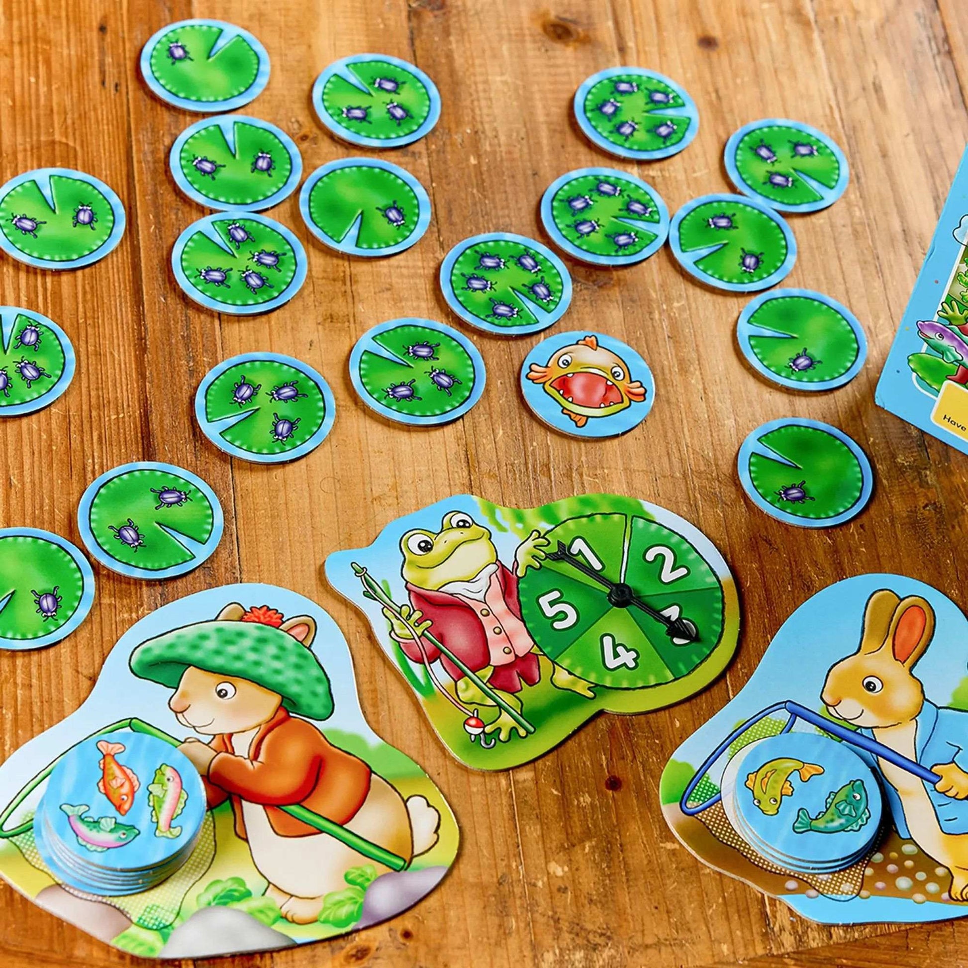 Orchard Peter Rabbit - Fish And Count game pieces including lily pads with bugs, a frog with a spinner, and rabbits fishing for fish tokens on a wooden table