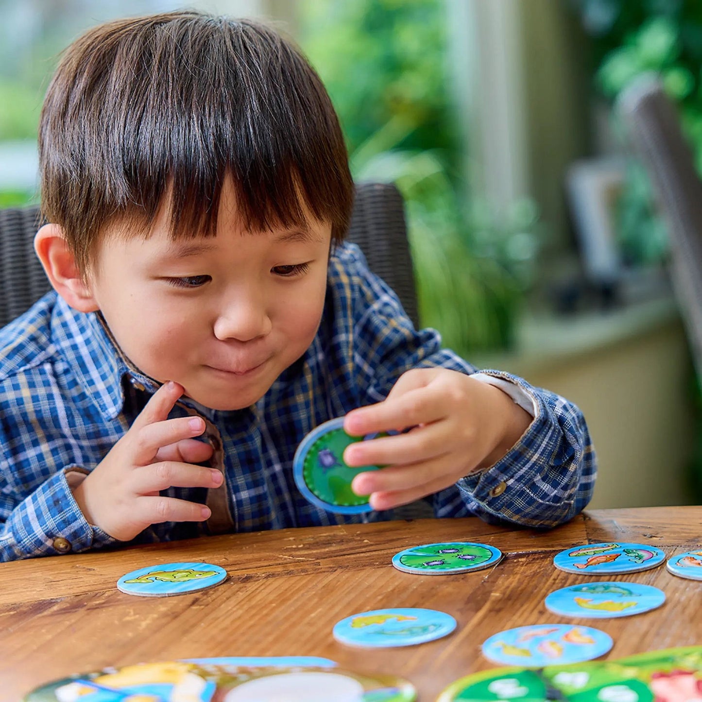 A young boy in a blue plaid shirt plays the Orchard Peter Rabbit Fish And Count game, holding a piece over a wooden table with more pieces