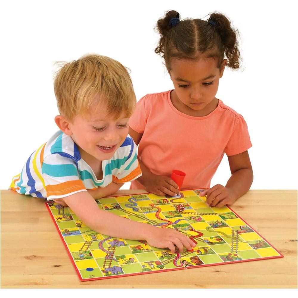 A boy and girl play a colorful Snakes and Ladders board game on a wooden table, the boy smiles moving a piece while the girl watches with a dice shaker