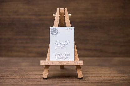 A white card on a small wooden easel displays an illustration of two pinkie fingers touching, with the text 9 PINKIES, all set against a dark wooden background