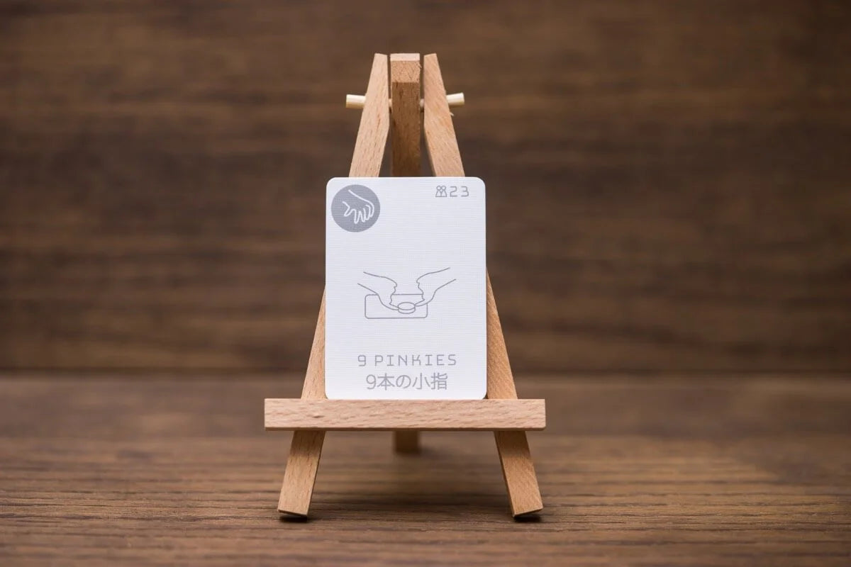 A white card on a small wooden easel displays an illustration of two pinkie fingers touching, with the text 9 PINKIES, all set against a dark wooden background