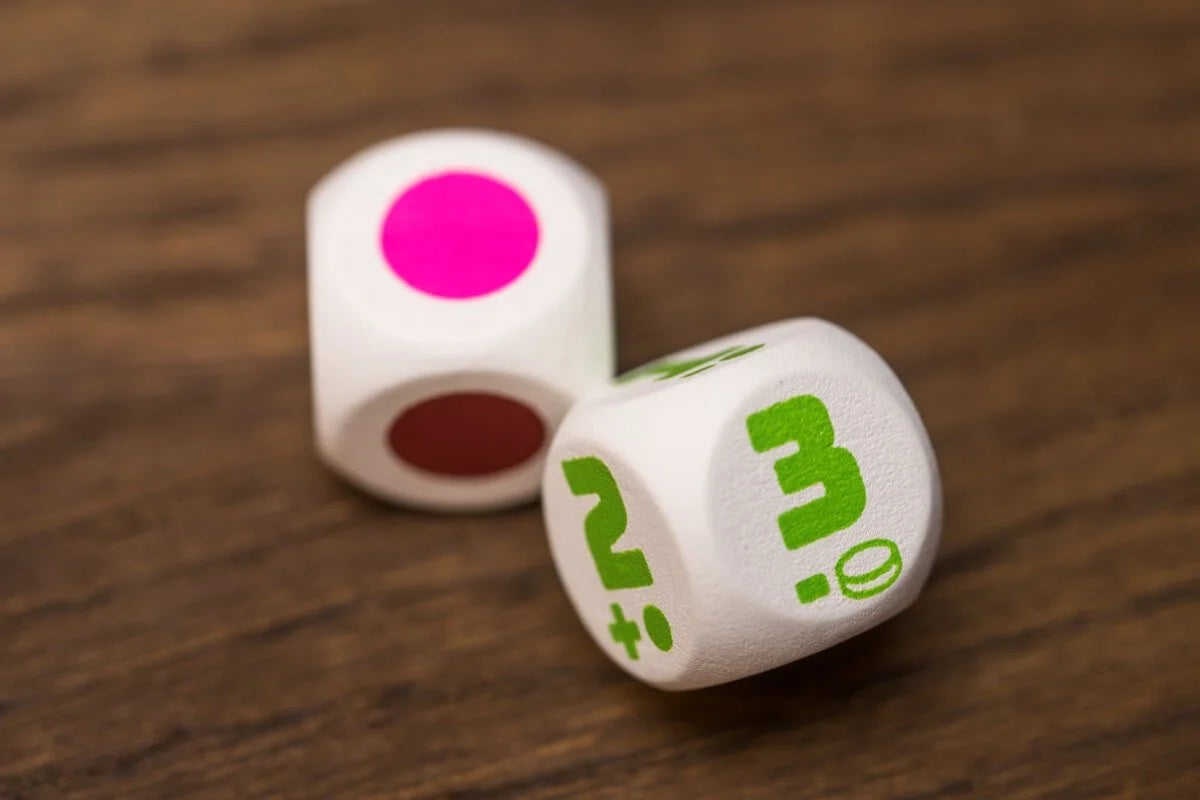 Two white dice rest on a wooden surface, one showing pink and red dots, the other displaying green numbers 2 and 3 with an icon