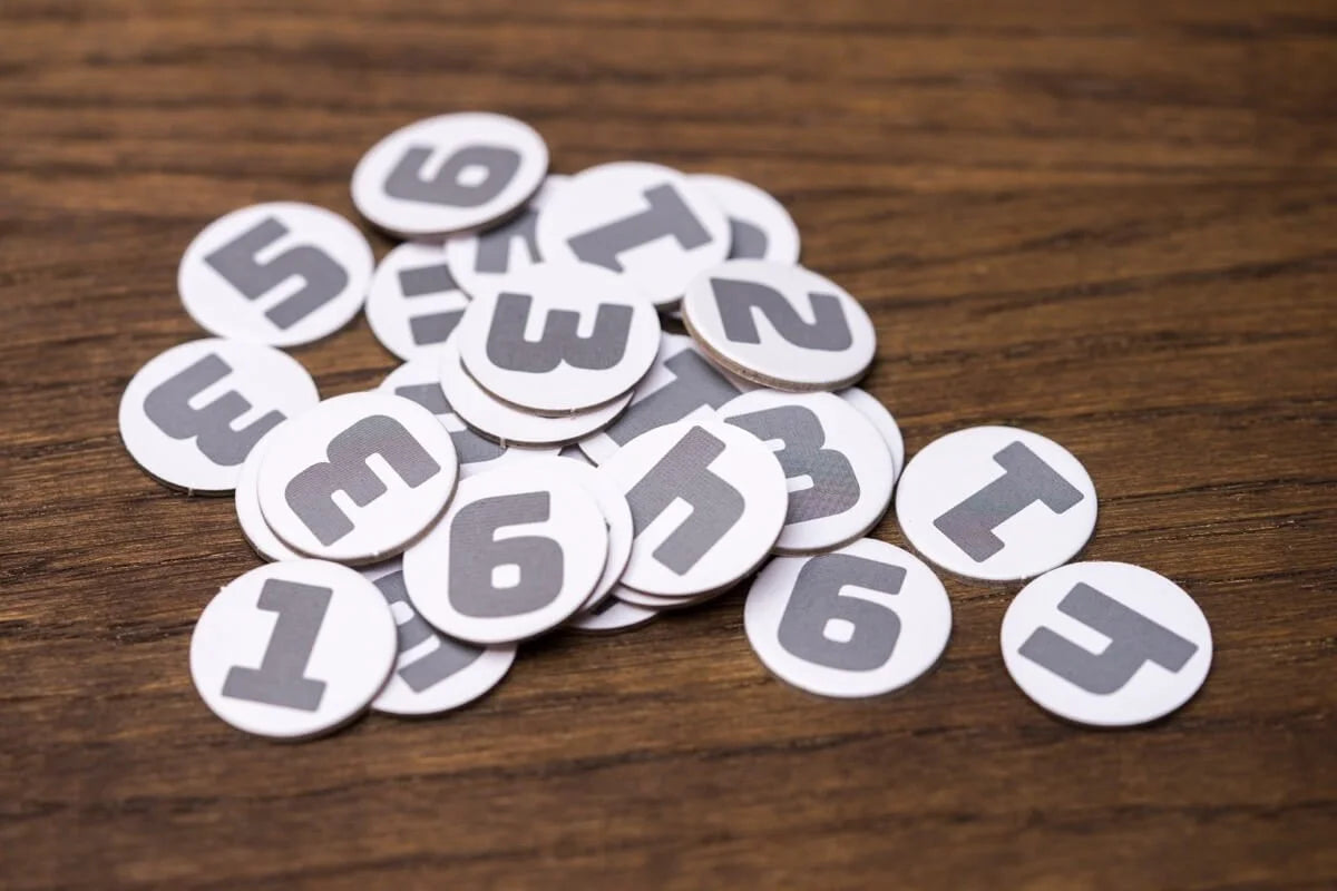 White round game tokens with grey numbers 1 through 6 are scattered on a brown wooden surface