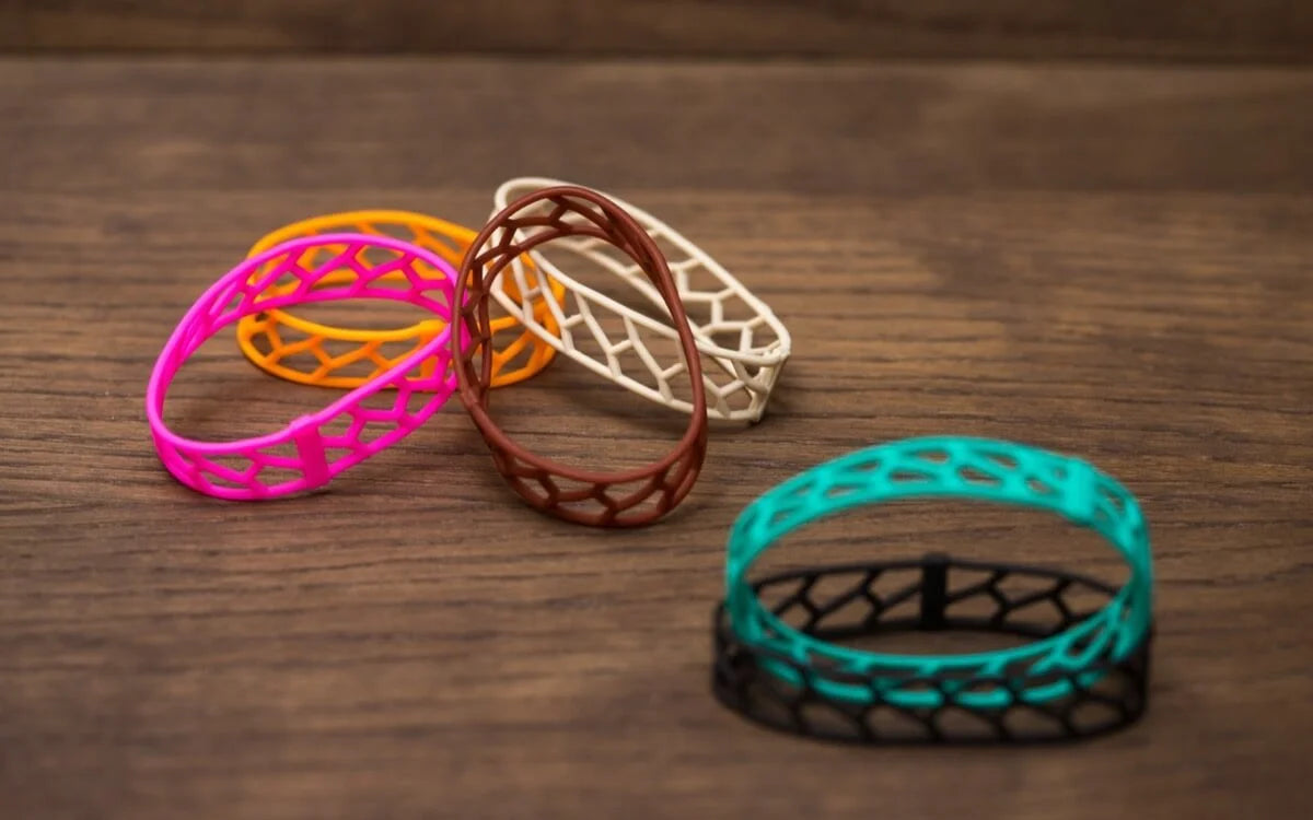 A group of colourful open-work wristbands in pink, orange, brown, white, and teal-over-black lay scattered on a wooden surface