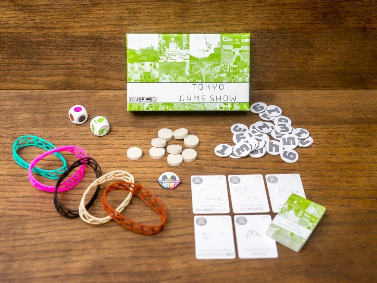 Tokyo Game Show board game components including its green box, two dice, colourful rubber bands, white and numbered grey tokens, and various game cards spread on a wooden table