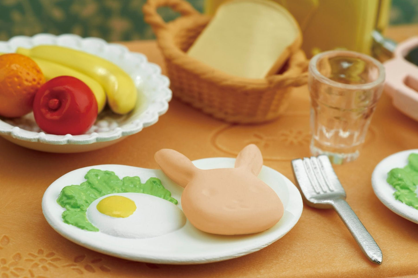 Miniature toy breakfast playset: a plate with a bunny-shaped item, egg, and lettuce, alongside fruit and bread