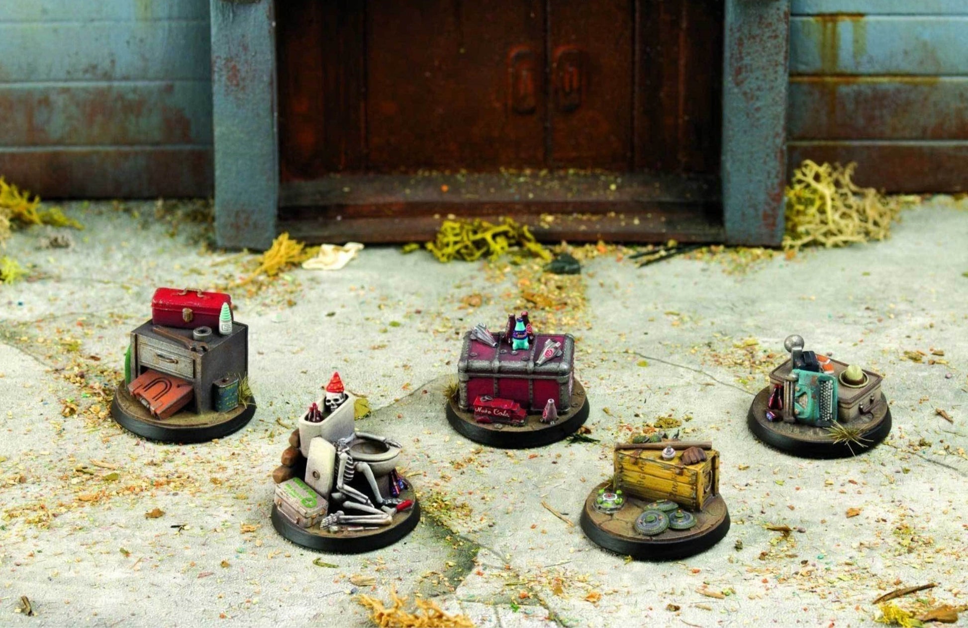Five detailed tabletop gaming terrain miniatures are scattered on a gritty ground in front of a weathered building, depicting various junk piles and a skeleton in a toilet