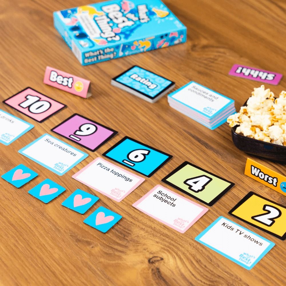 The What's the Best Thing card game is spread on a wooden surface showing number cards, topic prompts like "School subjects," heart tokens, and the blue game box behind a small bowl of popcorn
