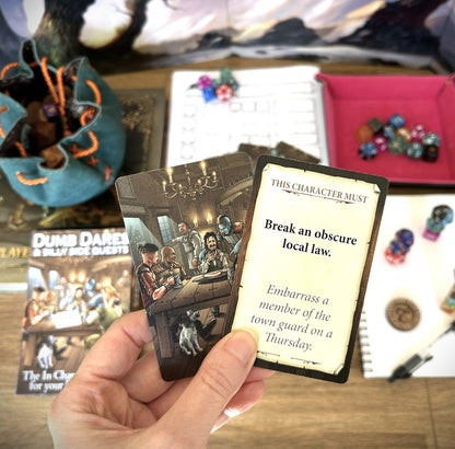 Hand holds two RPG quest cards; one shows a tavern scene, the other reads: "Break an obscure local law." Dice and books surround them