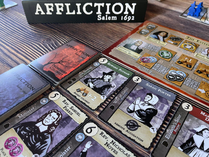 Tabletop view of the "Affliction Salem 1692" board game, showing character cards, action tokens, and blue player pieces