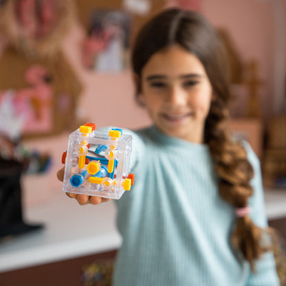A smiling girl holds up a clear plastic Criss Cross Cube maze featuring internal yellow, blue, and red puzzle tracks