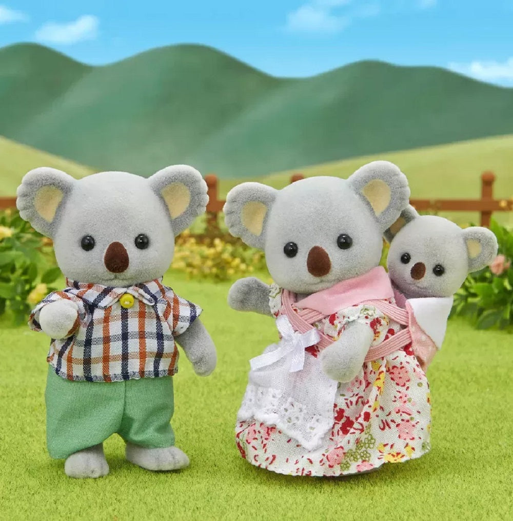 Flocked koala family figures (mom carrying baby, dad) standing on grass against a background of green hills