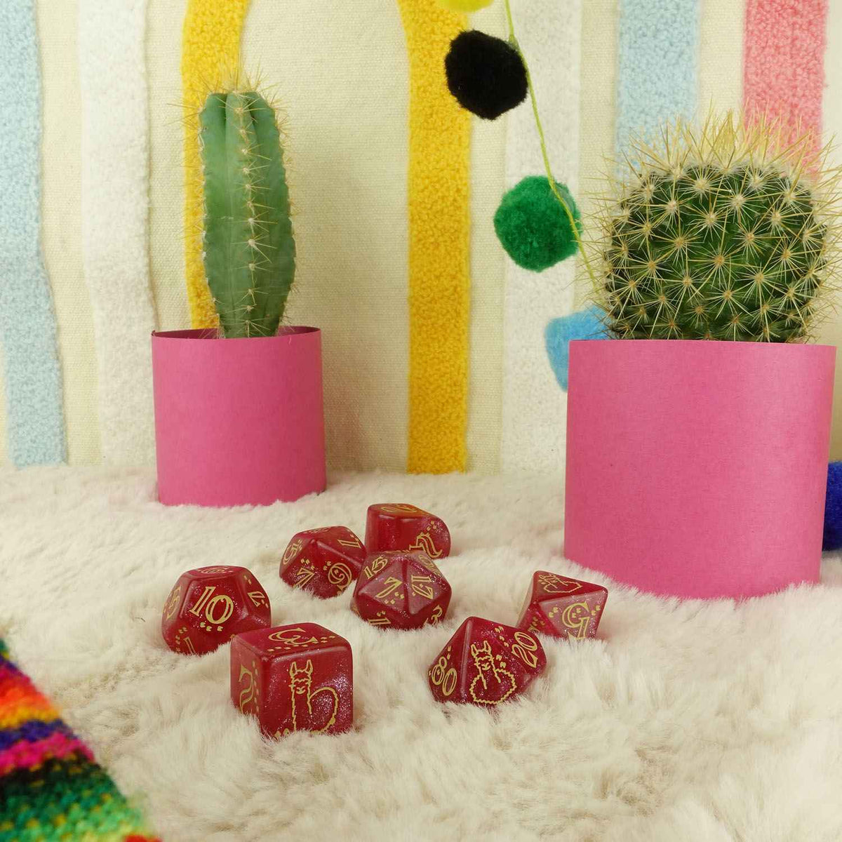 Red dice with gold markings scattered on a white furry rug, flanked by two cacti in pink pots. Colorful striped background.