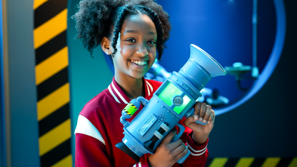 A smiling girl in a science lab setting holds the large blue Moose Despicable Me 4 Ultimate Fart Blaster with a glowing green reservoir and caution-striped accent