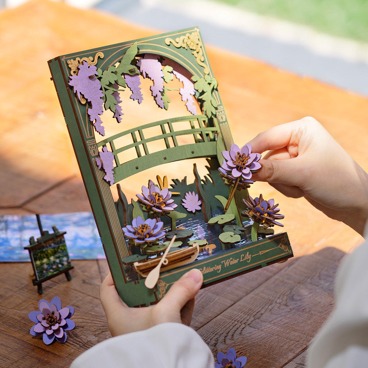 Hands assembling a wooden 3D puzzle of a garden scene with a bridge and purple water lilies