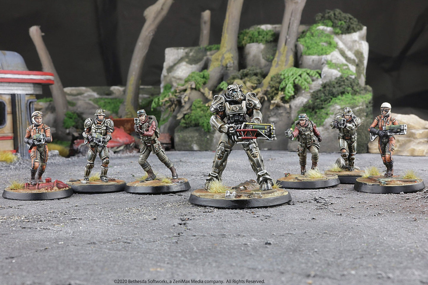 Seven painted Fallout miniatures, including a central Power Armor figure with a large weapon, stand armed on a textured battlefield