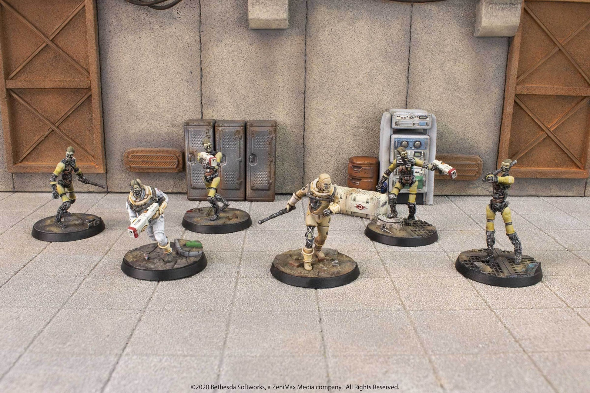 Six armed, ghoulish Fallout miniatures in a post-apocalyptic industrial scene, one wearing a white hazmat suit