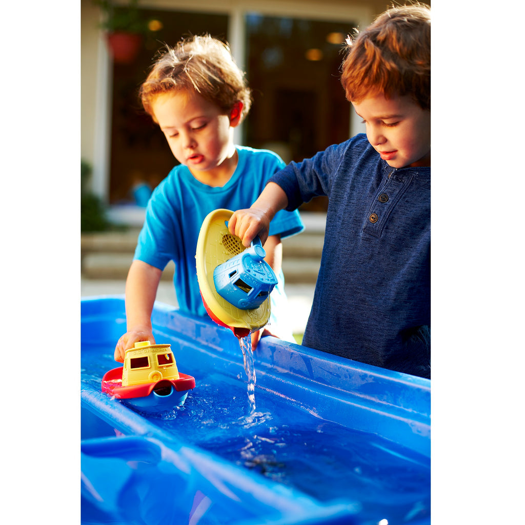 Two young boys pour water and float toy boats in a bright blue outdoor water table