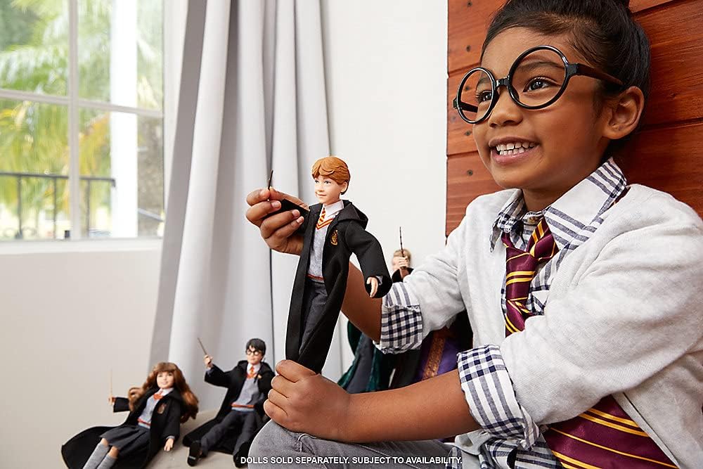 A smiling child with round glasses and a Gryffindor tie holds a Ron Weasley doll. Other Harry Potter dolls are behind