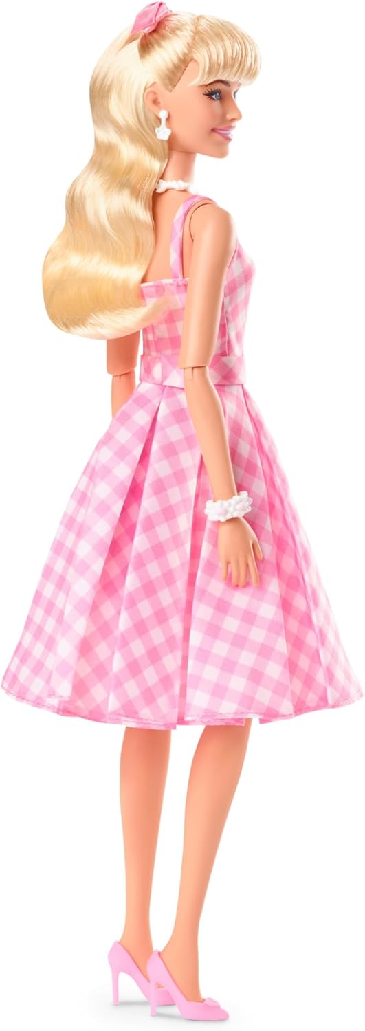 A blonde Barbie doll stands -  wearing a pink gingham dress -  pink high heels -  and a matching pink hair bow