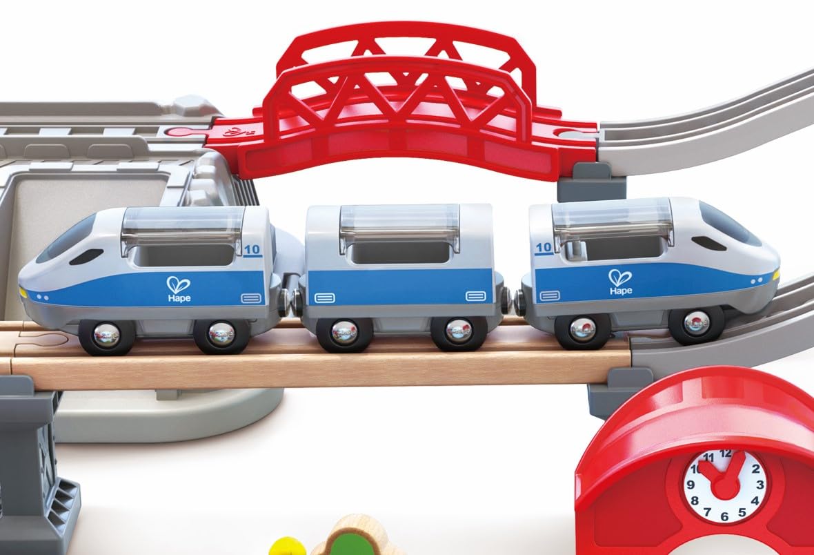 Blue and white Hape toy train traveling on a wooden bridge under a red plastic railroad structure
