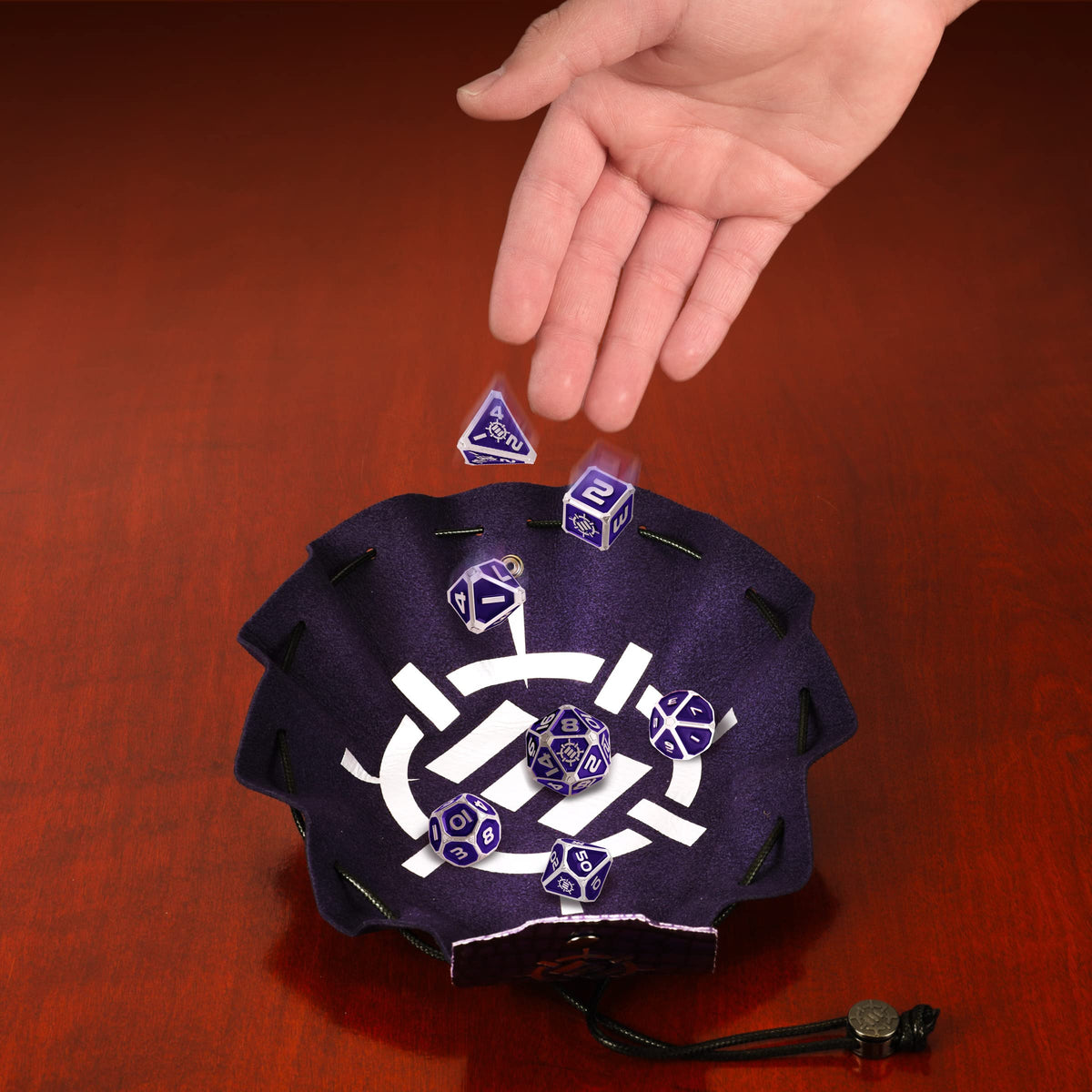 A hand drops purple metal RPG dice, some blurring from motion, into a purple vegan leather bag and tray with a white symbol on a wooden surface