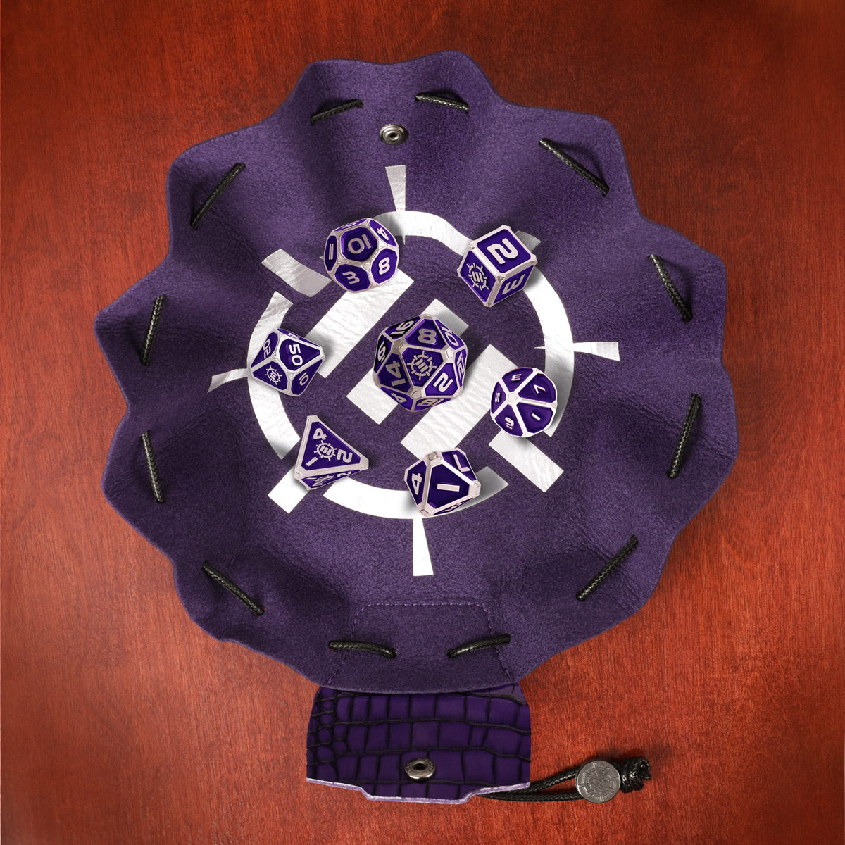 A purple 2-in-1 Vegan Leather Bag and Tray, featuring a white symbol, holds a Tabletop Collector&#39;s Metal RPG Dice Set in purple and silver, along with a snap closure and token on wood