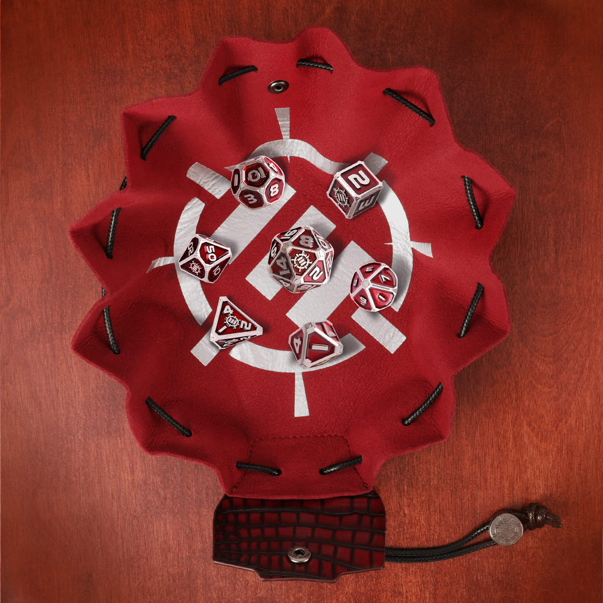 A red vegan leather dice bag and tray with a white design, displaying a metal RPG dice set in red and silver on a wooden background