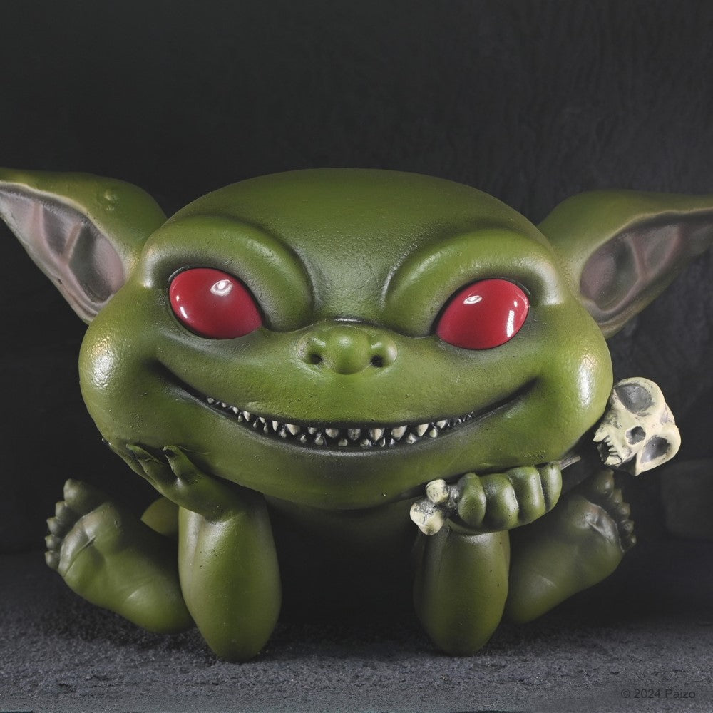 A green baby goblin with red eyes and a toothy grin rests its chin on its hand, holding a small skull on a stick