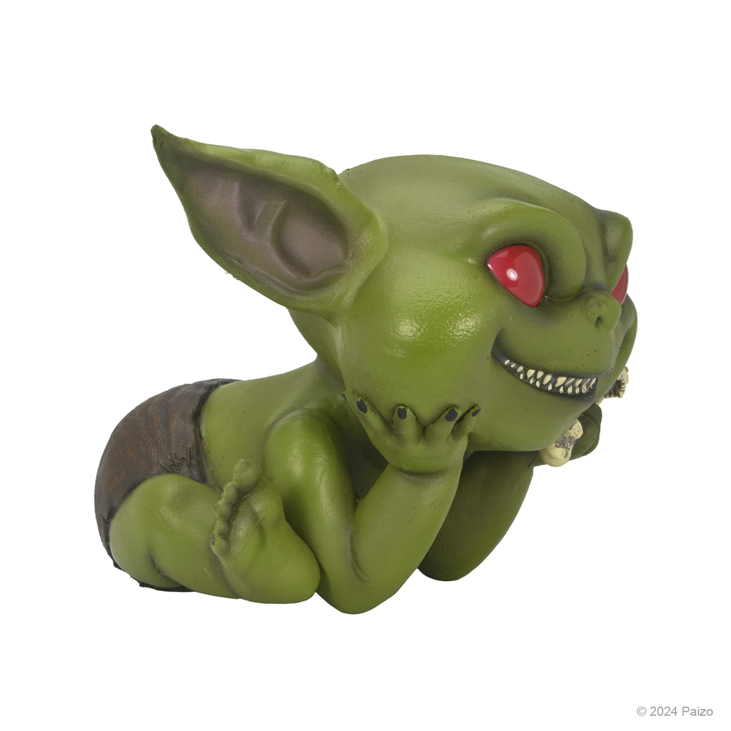 Mischievous green baby goblin figure with red eyes, sharp teeth, lying on its stomach with head resting on hands