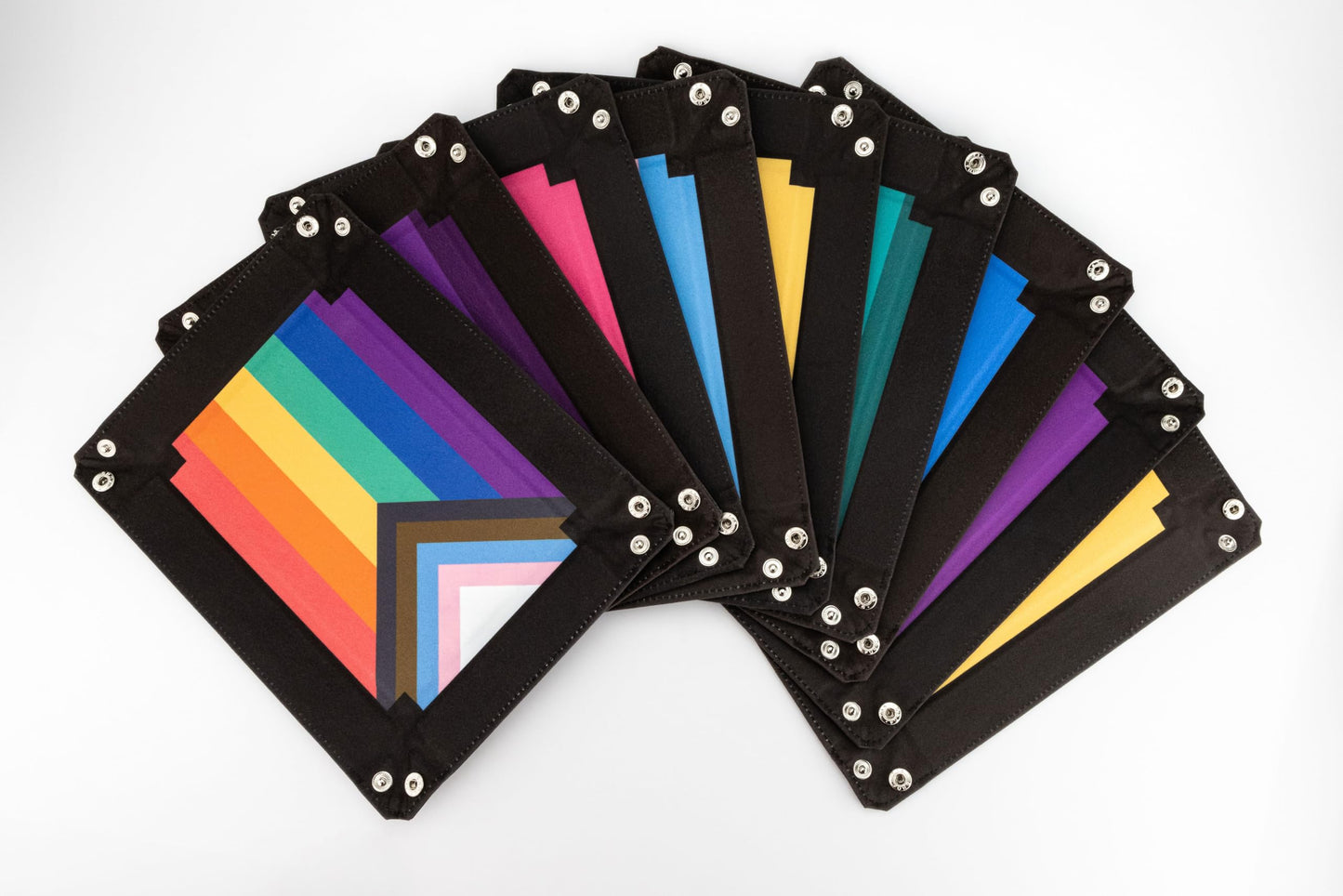 A fan of black velvet foldable dice trays on a white background, featuring the Progress Pride flag design and other vibrant flag colors with silver snaps