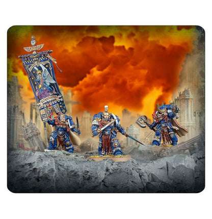 Three Ultramarines miniatures holding weapons and a large banner stand on ruins beneath a fiery orange sky