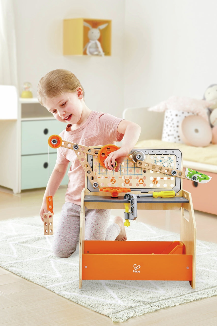A smiling child kneels on a rug playing with the Hape Discovery Scientific Workbench, manipulating its wooden crane and orange plastic gears