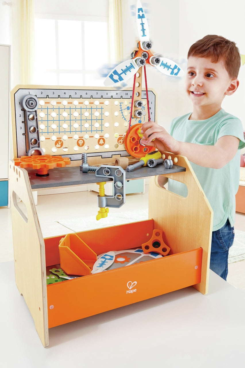 Happy boy assembling pieces on the wooden Hape workbench, actively turning a pulley that operates the attached propeller
