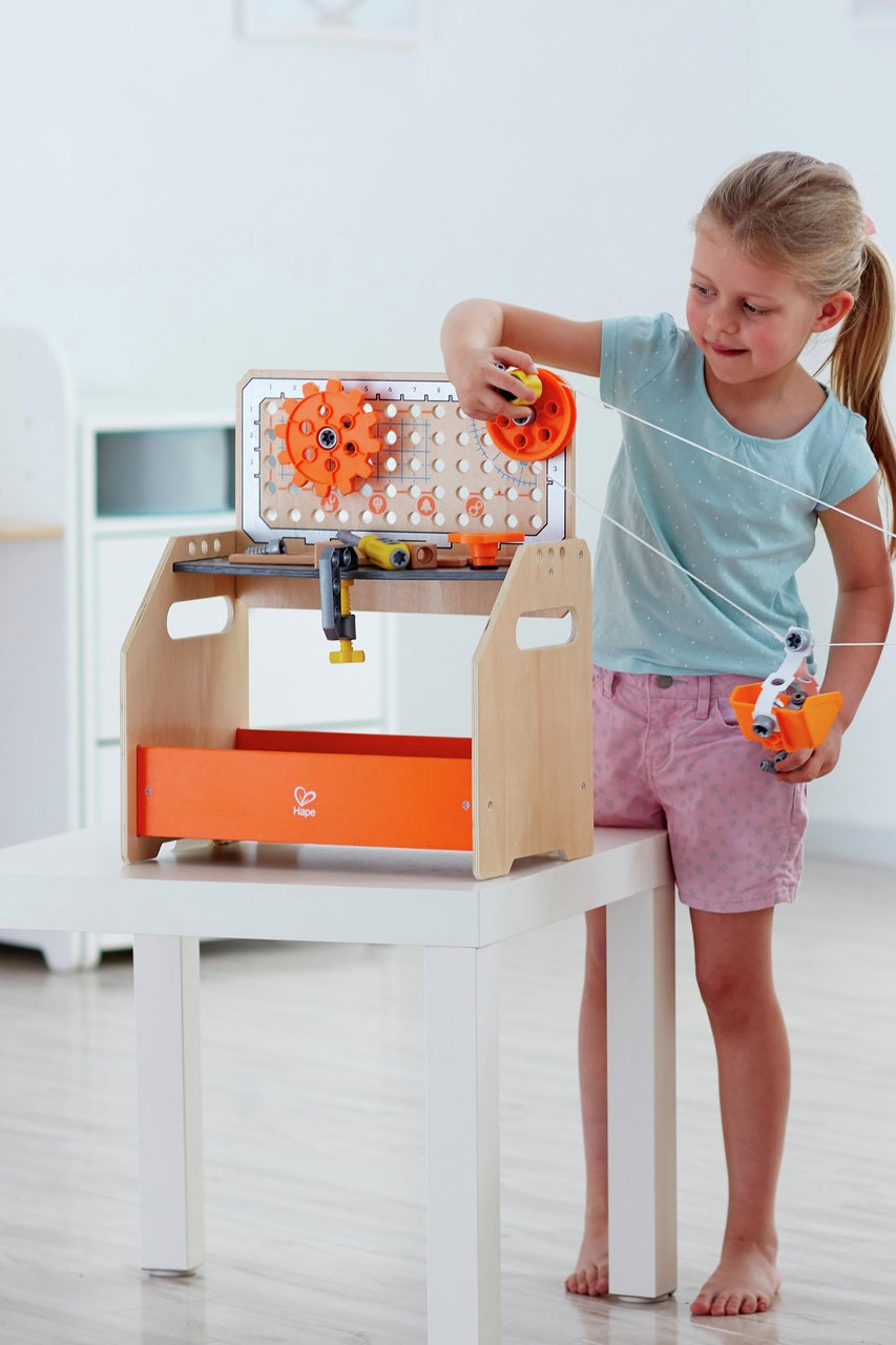 A smiling young girl operates the wooden Hape Discovery Scientific Workbench with its visible orange gears, pegboard back, and string pulley system on a white surface